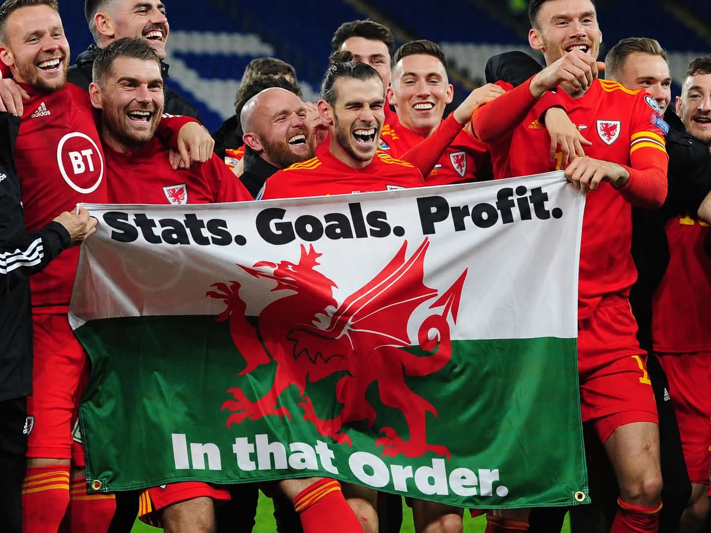 Wales football players celebrating with a banner reading Stats. Goals. Profit. In that Order.